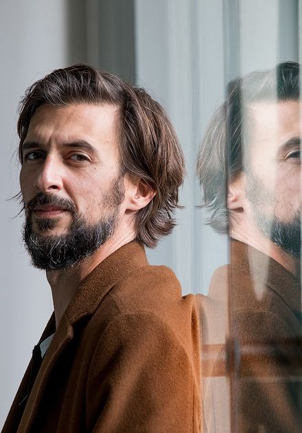 A man with brown hair and a beard in a brown coat, with his reflection visible in a glass surface.